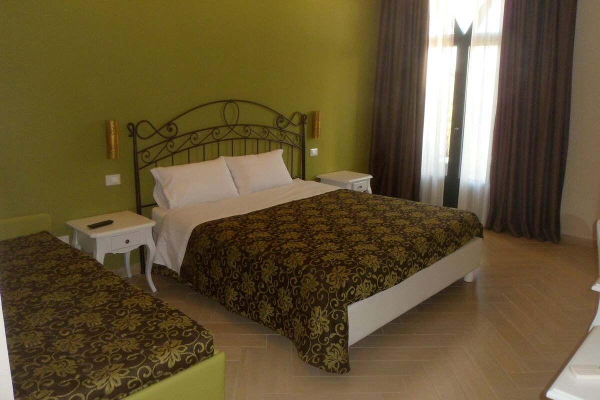 Une chambre d’hôtel avec un lit, deux tables de chevet, une commode et une fenêtre avec des rideaux.