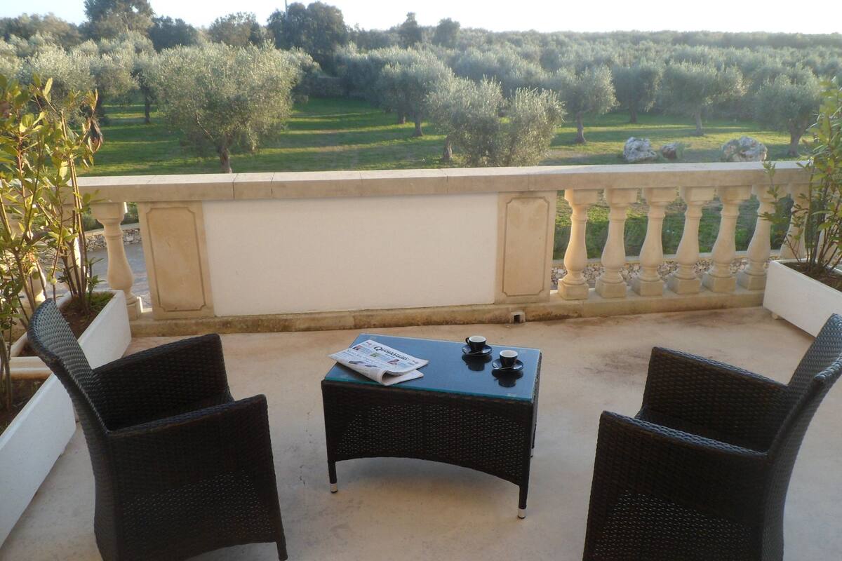 Un balcon agrémenté de meubles en osier, une table avec un journal et une vue sur des oliviers et la verdure environnante.