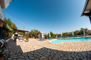 A stone-floored outdoor pool area with lounge chairs and umbrellas, a barbecue setup, and a pergola. - Apartments Ithaki Holidays (Lefkada)