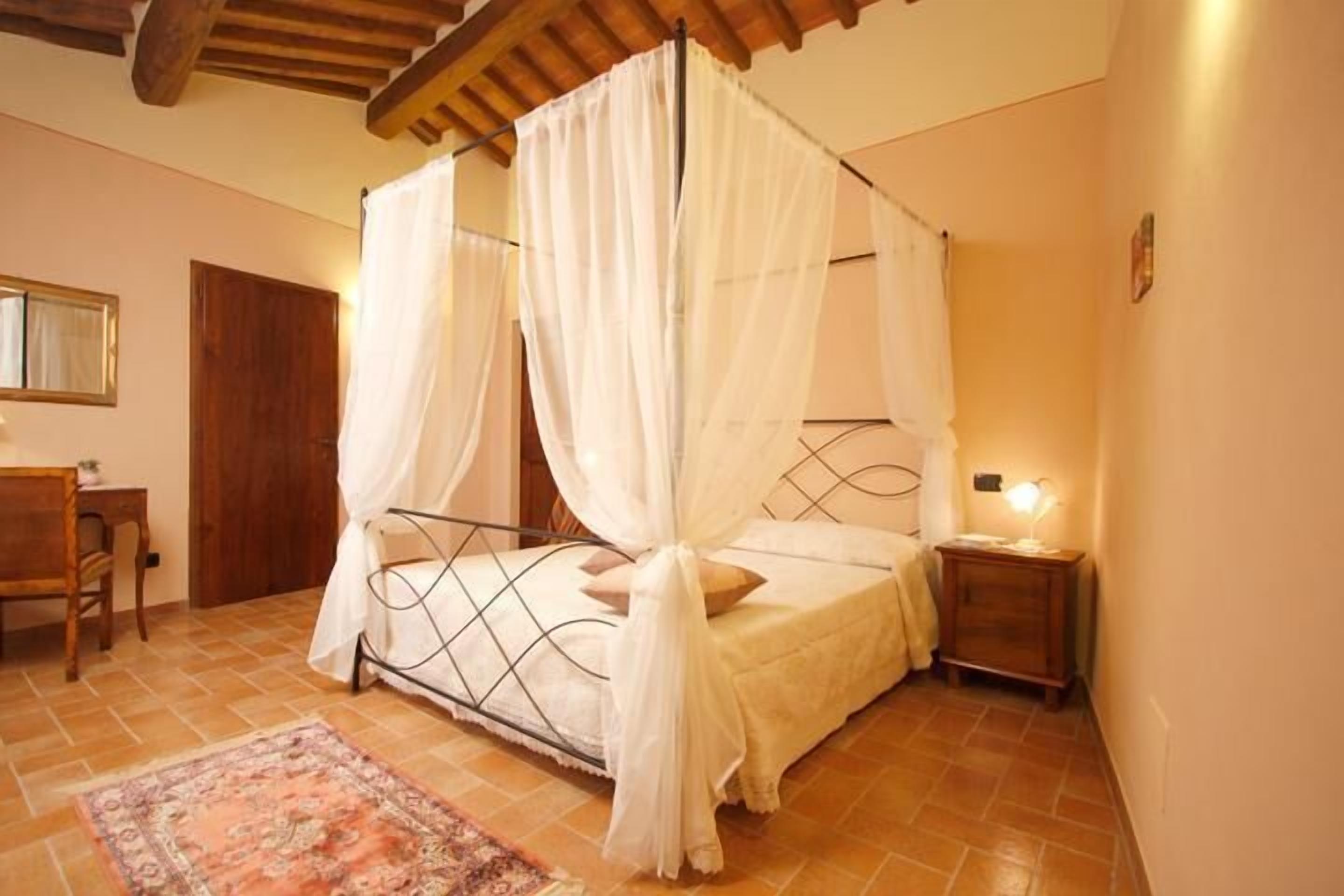 A bedroom with a four-poster bed, a wooden desk, a chair, and a nightstand with a lamp.