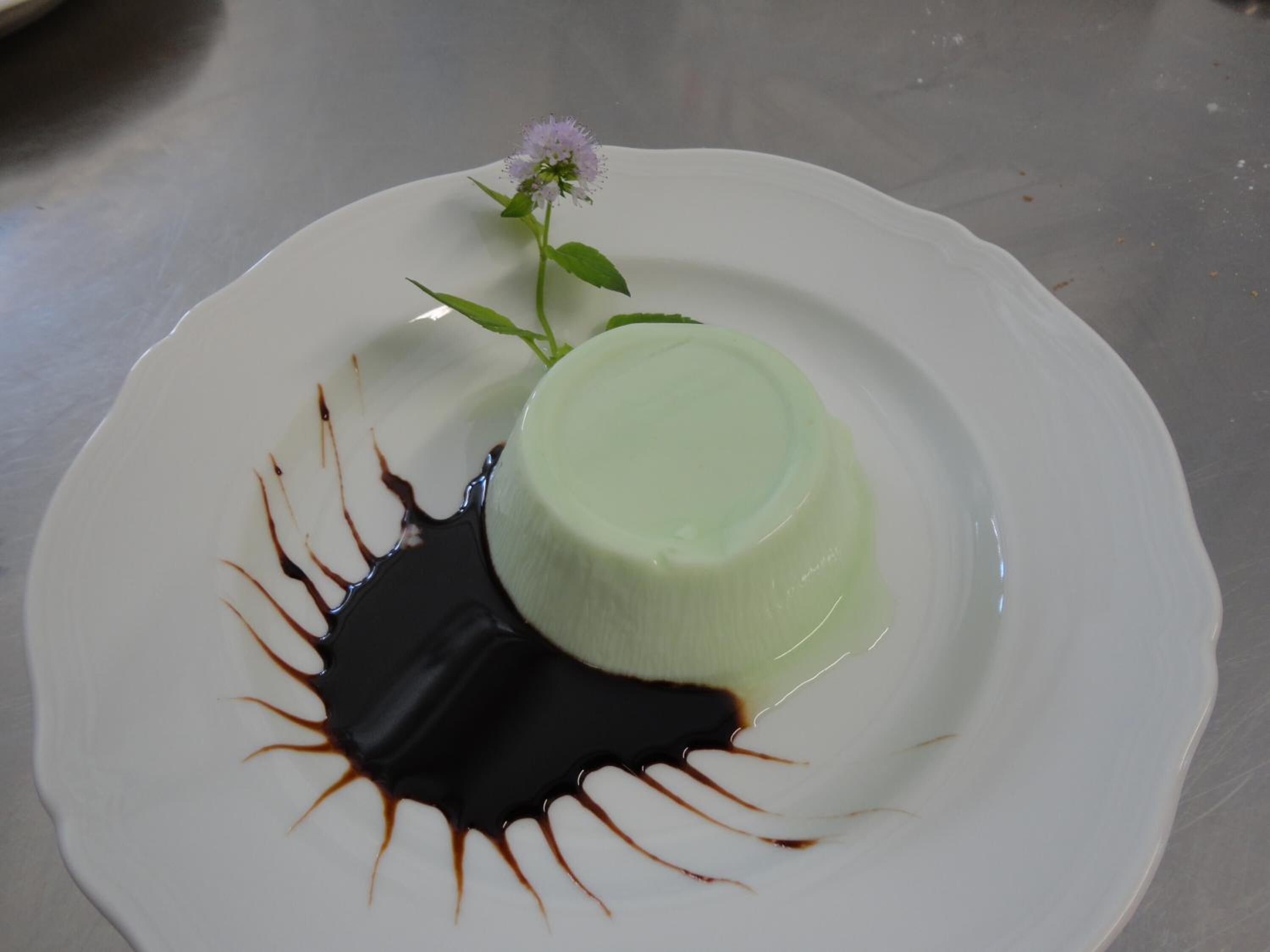 A dessert plate with a panna cotta and chocolate sauce, accompanied by a small flower.
