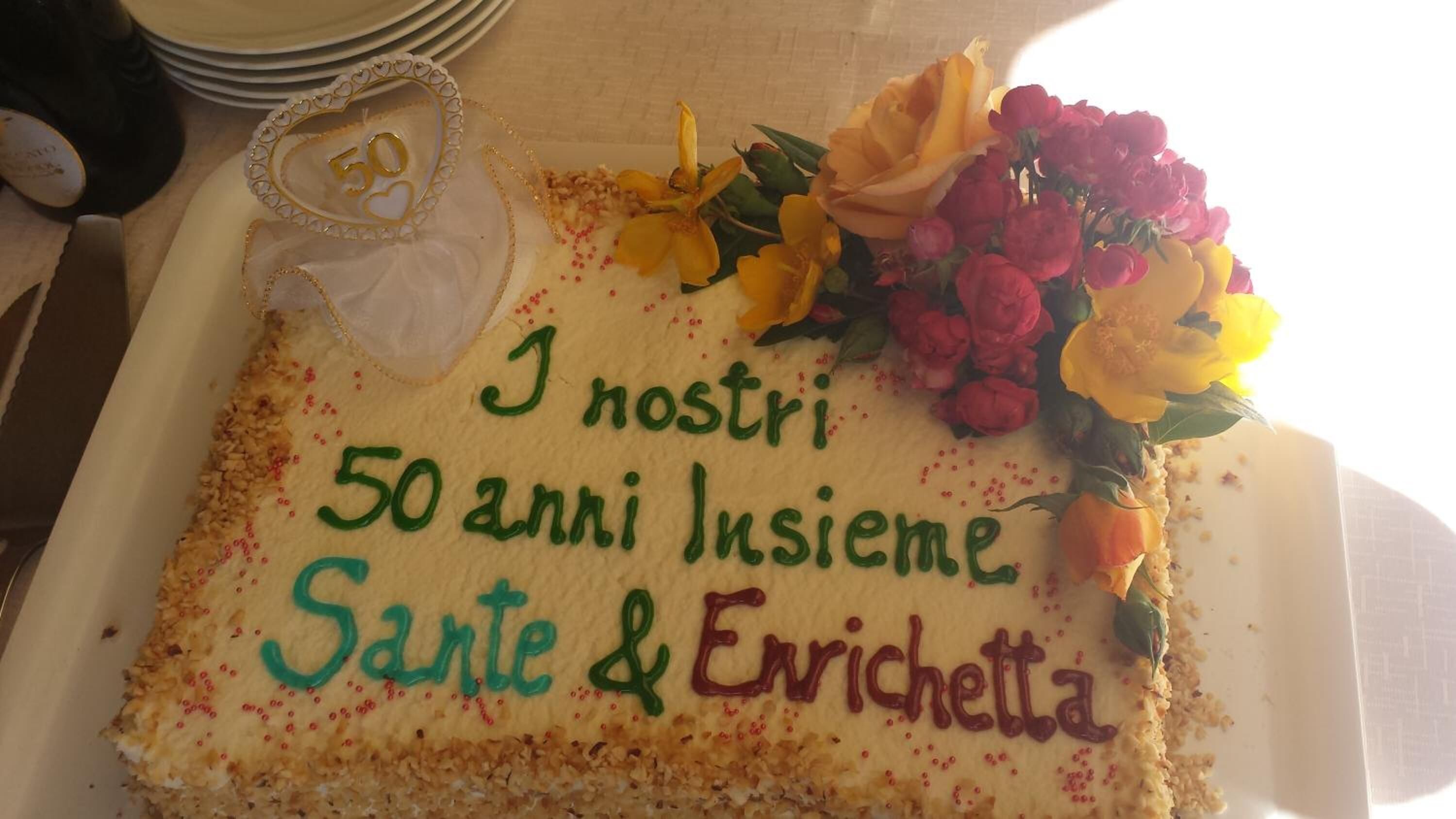A cake with "I nostri 50 anni insieme Sante & Evichetta" written on it, decorated with flowers and a heart-shaped plate.