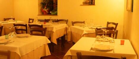 Un restaurante con varias mesas, cada una con mantel blanco, sillas de madera y suelo de baldosas.