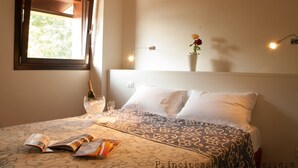 Una cama bien hecha con una colcha estampada, una mesita de noche con una botella de champán, copas y una revista. Se ve un ventanal con vistas a zonas verdes.