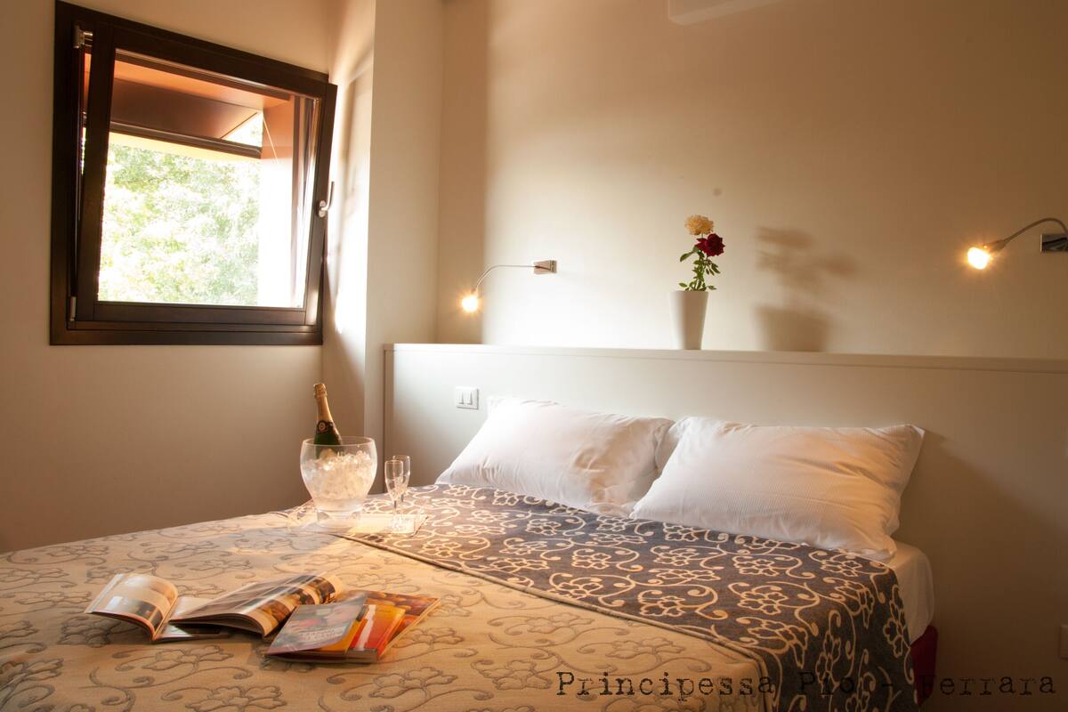 Una cama bien hecha con una colcha estampada, una mesita de noche con una botella de champán, copas y una revista. Se ve un ventanal con vistas a zonas verdes.