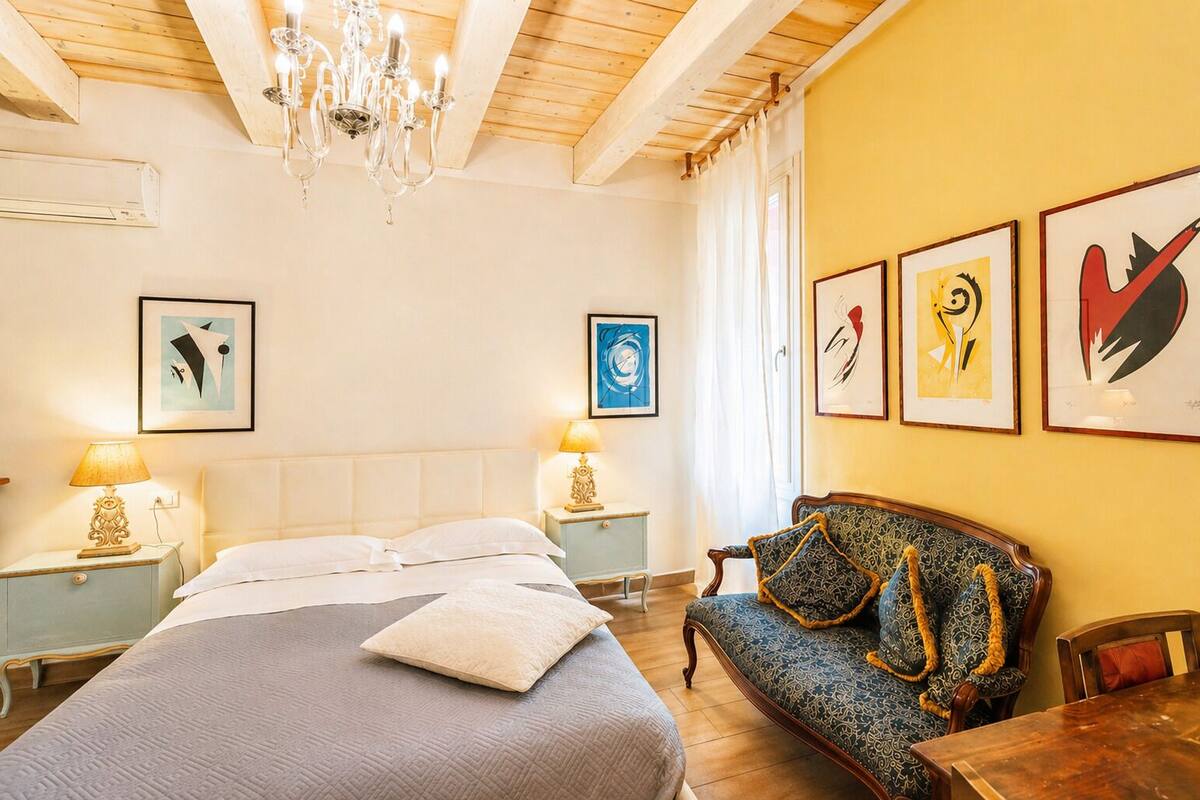 A bedroom with a bed, bedside tables, a chandelier, artwork on the walls, and a sofa.