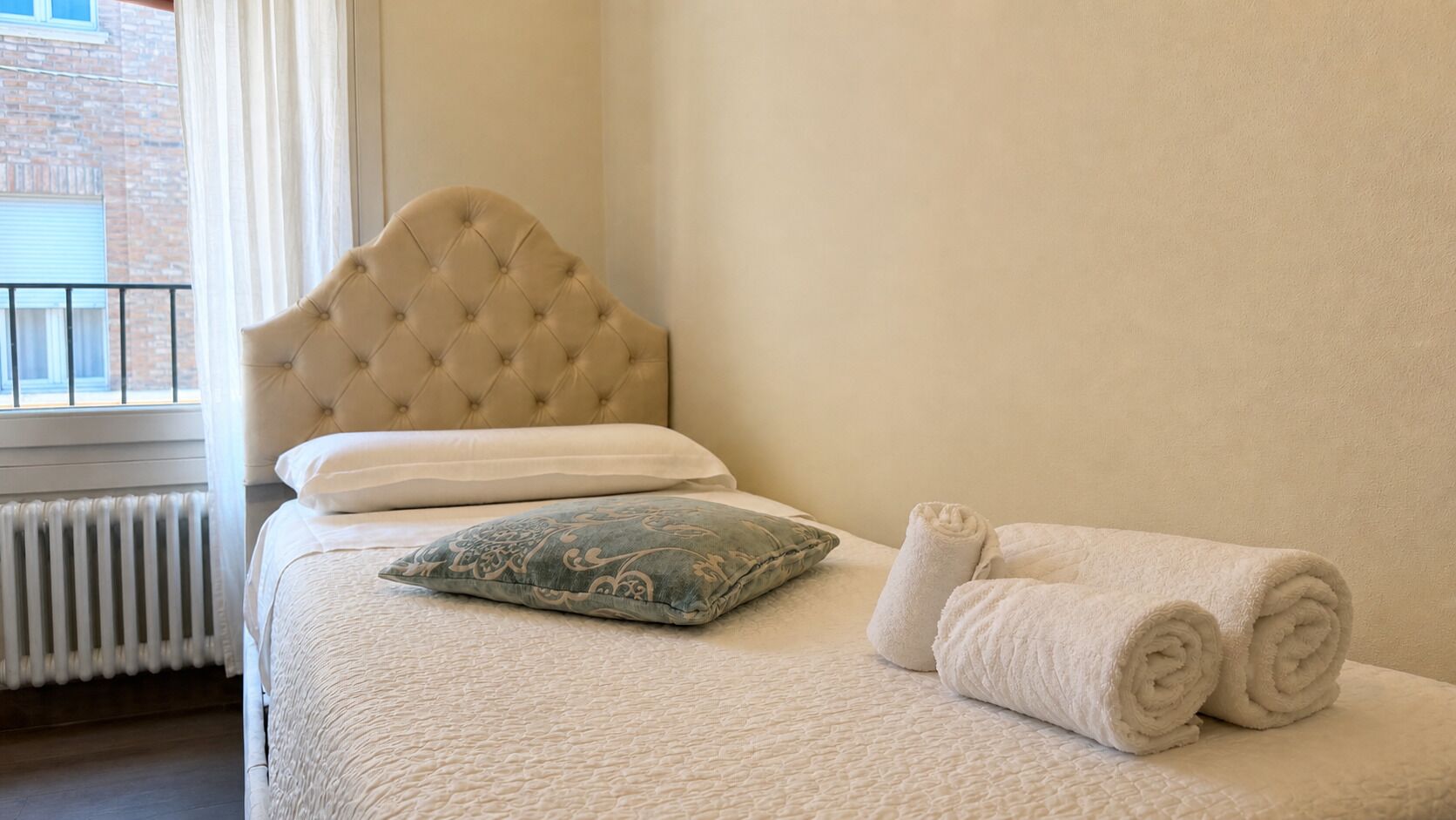 A neatly made bed with a tufted headboard, a patterned pillow, and rolled towels.