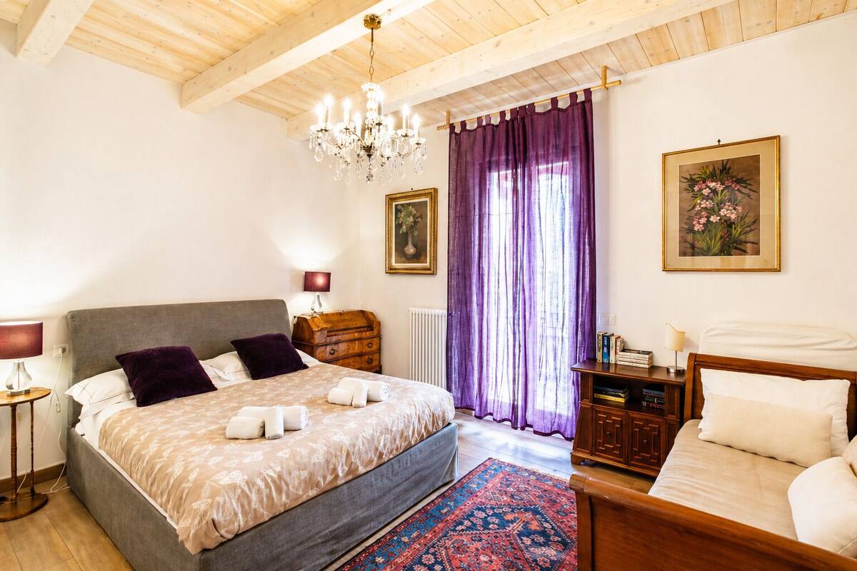 A bedroom with a large bed, a wooden headboard, a chandelier, a dresser, and two paintings on the wall.