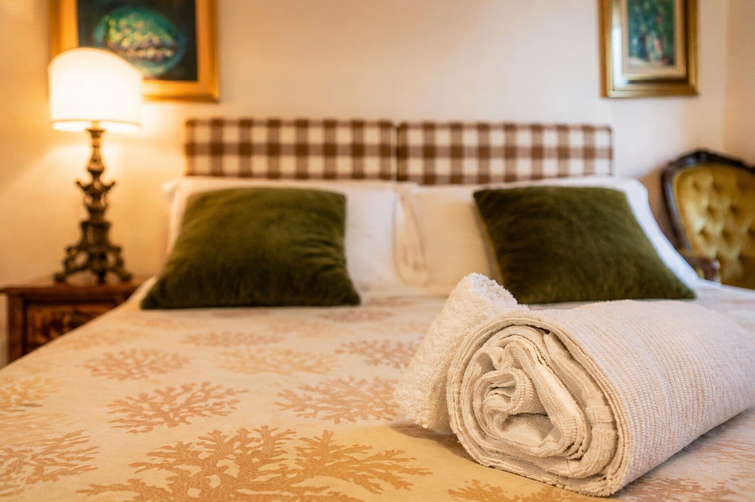 A neatly made bed with a checkered headboard, two pillows, a rolled towel, and a bedside lamp.