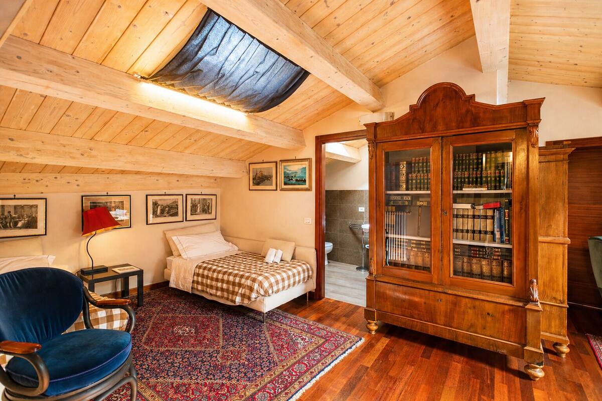 A bedroom with a bed, a blue chair, a red lamp, a wooden cabinet, and a skylight.