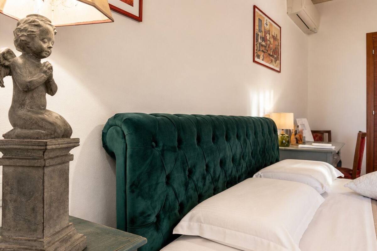 A hotel room with a green upholstered headboard, a statue on a pedestal, a bed with white linens, a wooden door, and a framed picture on the wall.