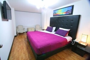 A bedroom with a large bed, a flat-screen TV mounted on the wall, a bedside table with a lamp, and a painting above the bed. - Hotel Lemus Plaza (Bogota)