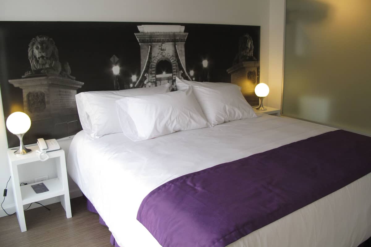 Habitación de hotel con cama, ropa de cama blanca, un plaid morado, una mesita de noche con lámpara y un mural en la pared.