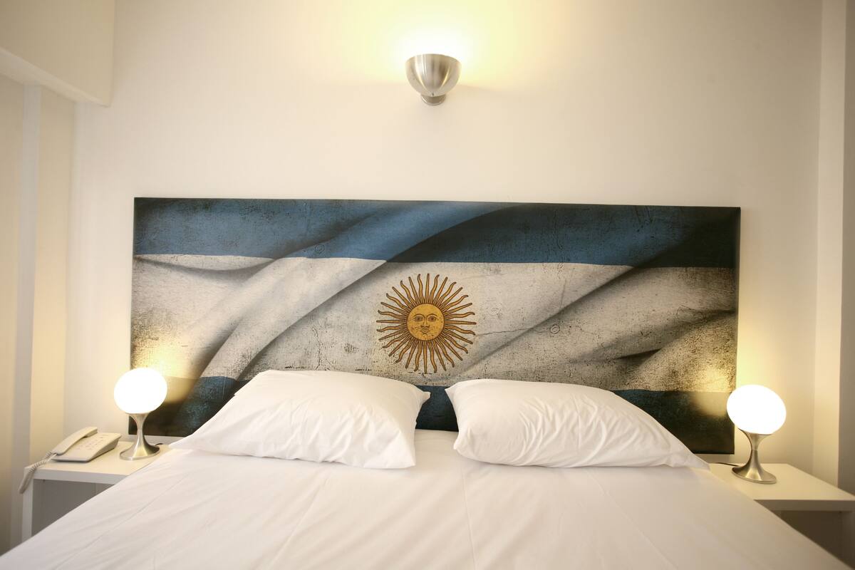 Una cama con sábanas blancas, dos lámparas de noche, una lámpara de pared y un cuadro abstracto grande con motivos de sol.