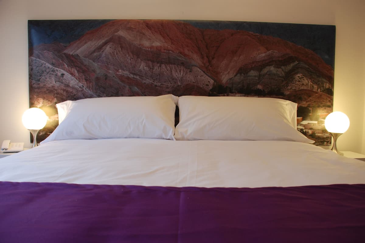 Una cama con sábanas blancas y una colcha morada, con dos lámparas de noche a cada lado.