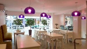 Un cafè modern amb taules i cadires blanques, llums penjants morades i plantes en testos.