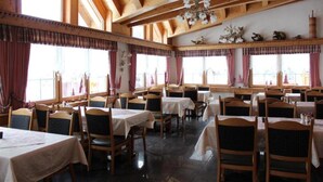 A dining area with wooden tables and chairs, large windows with curtains, and a chandelier. - Eden (SAAS GRUND)
