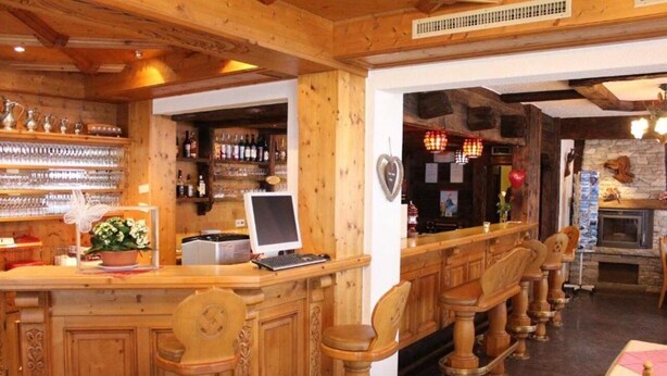 A wooden bar area with bar stools, a laptop on the counter, and shelves with bottles and glasses. - Eden (SAAS GRUND)