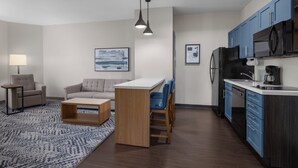 A modern living room with a sofa, coffee table, and a small kitchen area with a refrigerator, microwave, and cabinets. - Candlewood Suites Alexandria by IHG (Alexandria)