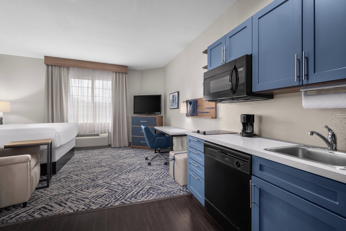 A hotel room with a bed, desk, kitchenette, and a TV.