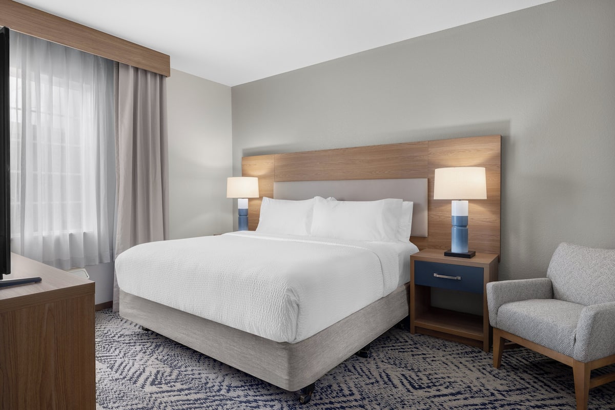 Una habitación de hotel con una cama grande, dos mesitas de noche con lámparas, una silla y un escritorio con televisor.