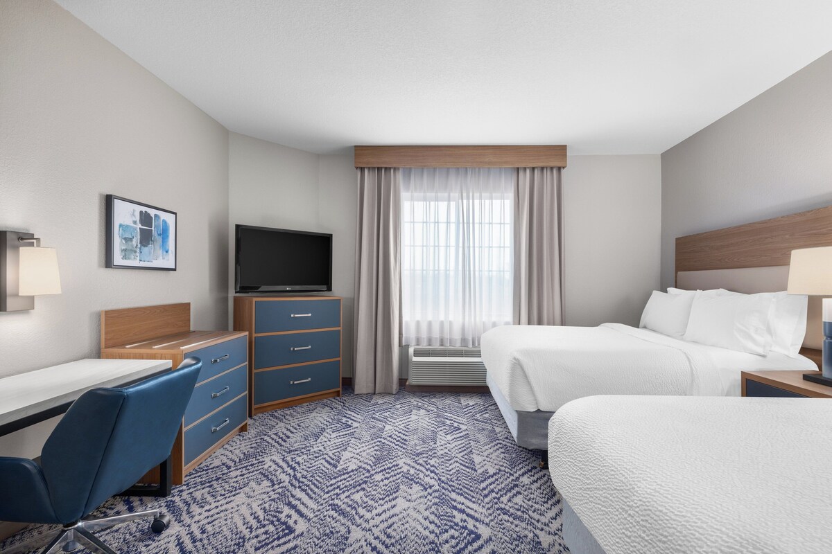 Ein Hotelzimmer mit Bett, Schreibtisch, Stuhl, Fernseher und einem Fenster mit Vorhängen.