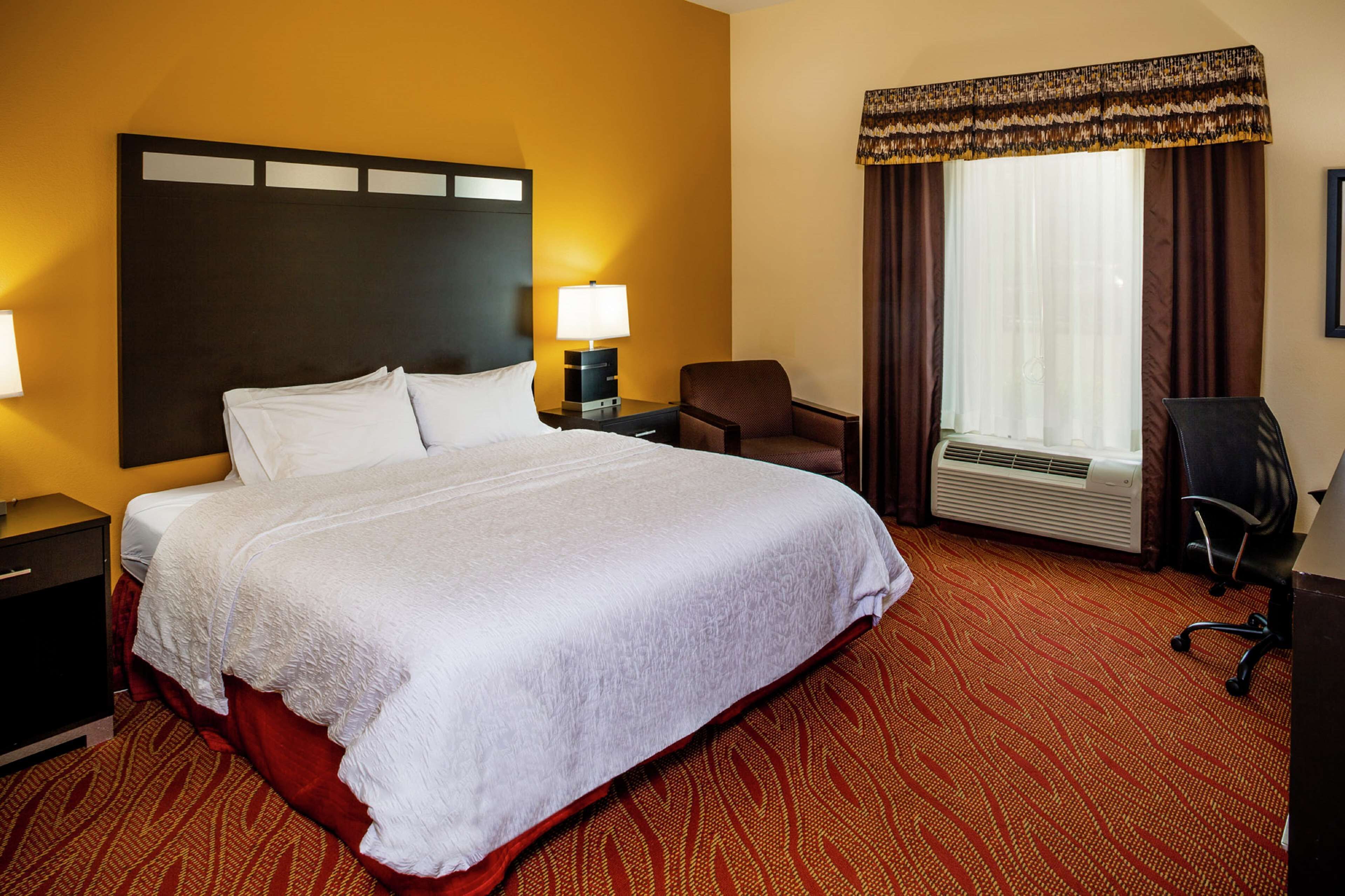 A hotel room with a large bed, two bedside tables with lamps, a chair, a desk, and a window with curtains.
