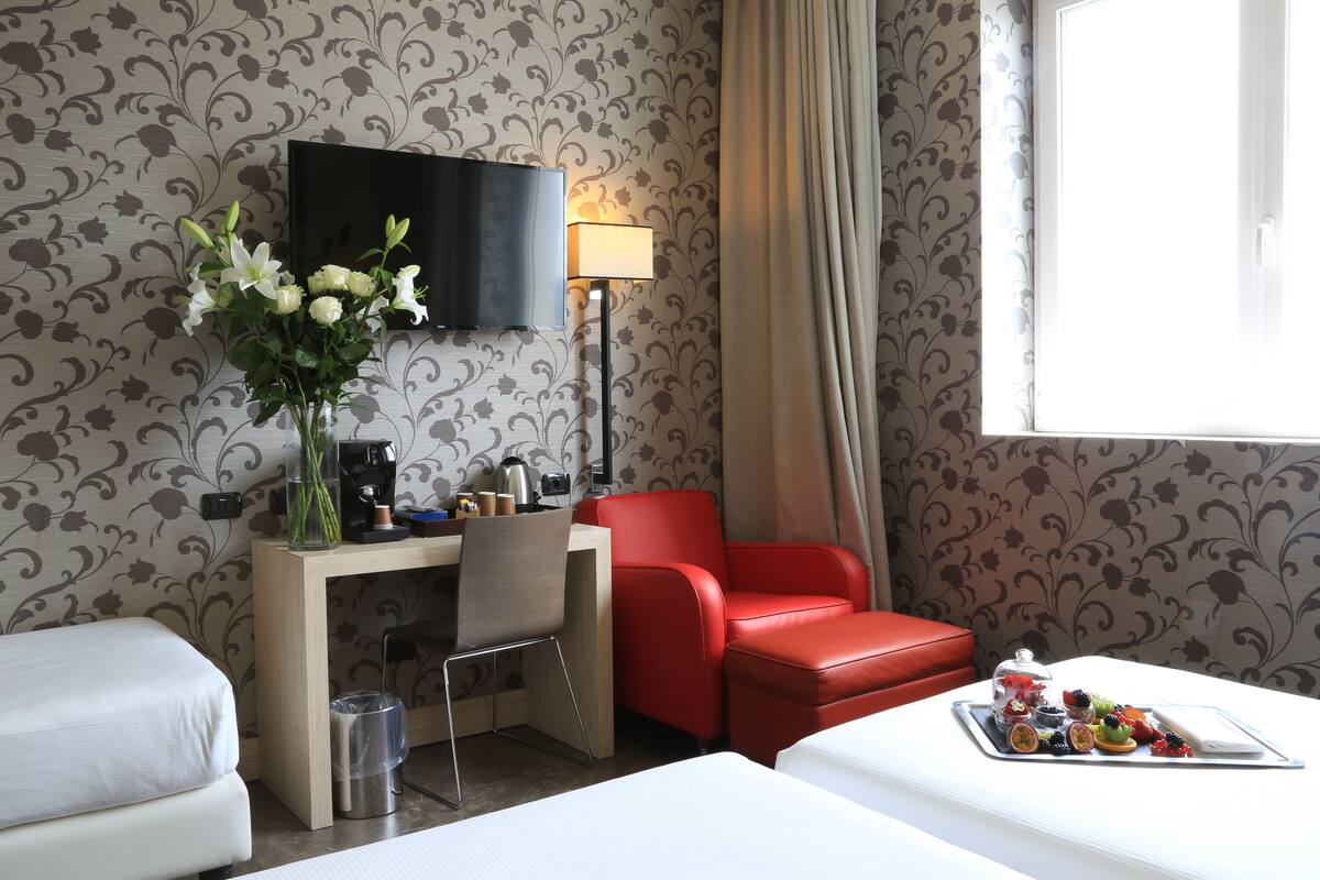A hotel room with a bed, a desk with a chair, a red armchair, a TV, a lamp, and a tray of fruit.