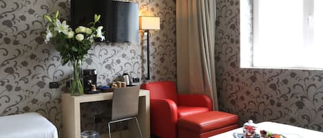 A hotel room with a bed, a desk with a chair, a red armchair, a TV, a lamp, and a tray of fruit.