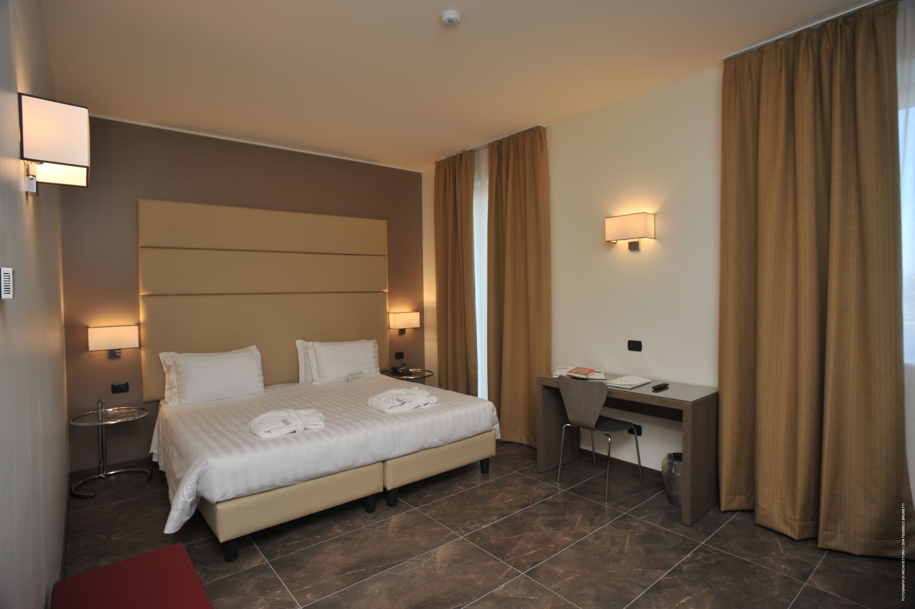 Quarto de hotel com cama, mesas de cabeceira, escrivaninha e uma cadeira.