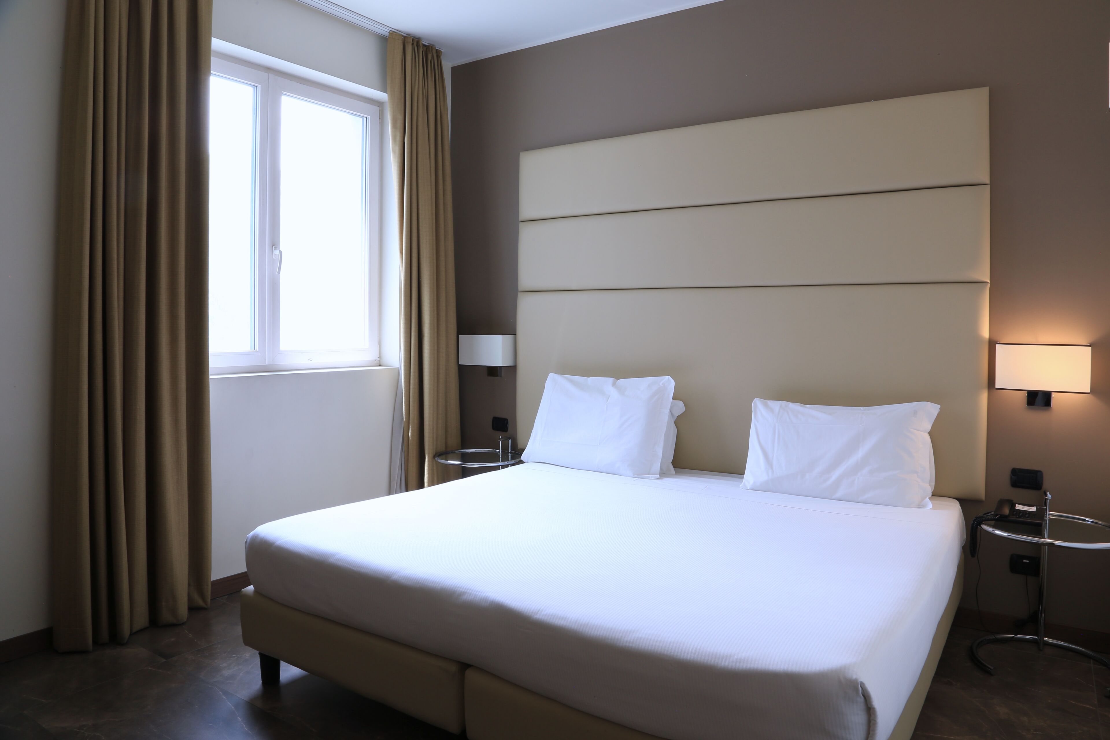 Habitació d'hotel amb un llit gran, dues taules de nit, una finestra amb cortines i una làmpada penjada a la paret.