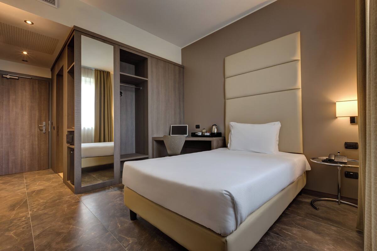 A modern hotel room with a large bed, a desk with a computer, a bedside table with a lamp, and a walk-in closet.