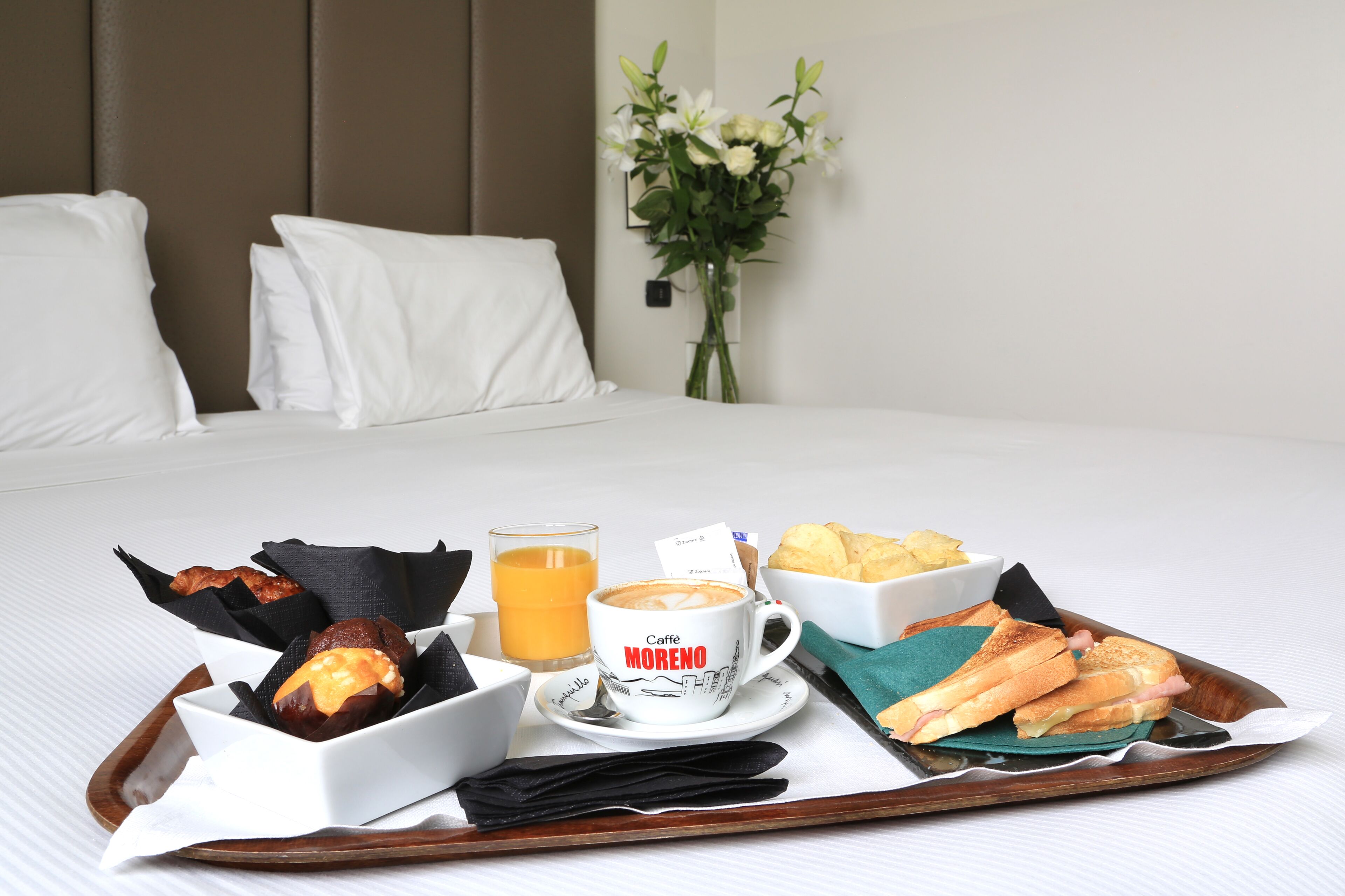A breakfast tray with croissants, a coffee cup labeled "Caffe MORENO", orange juice, and a sandwich on a bed with white bedding and pillows.