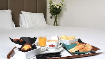 Bandeja de desayuno con cruasanes, una taza de café con la etiqueta "Caffe MORENO", zumo de naranja y un sándwich sobre una cama con ropa blanca y almohadas.