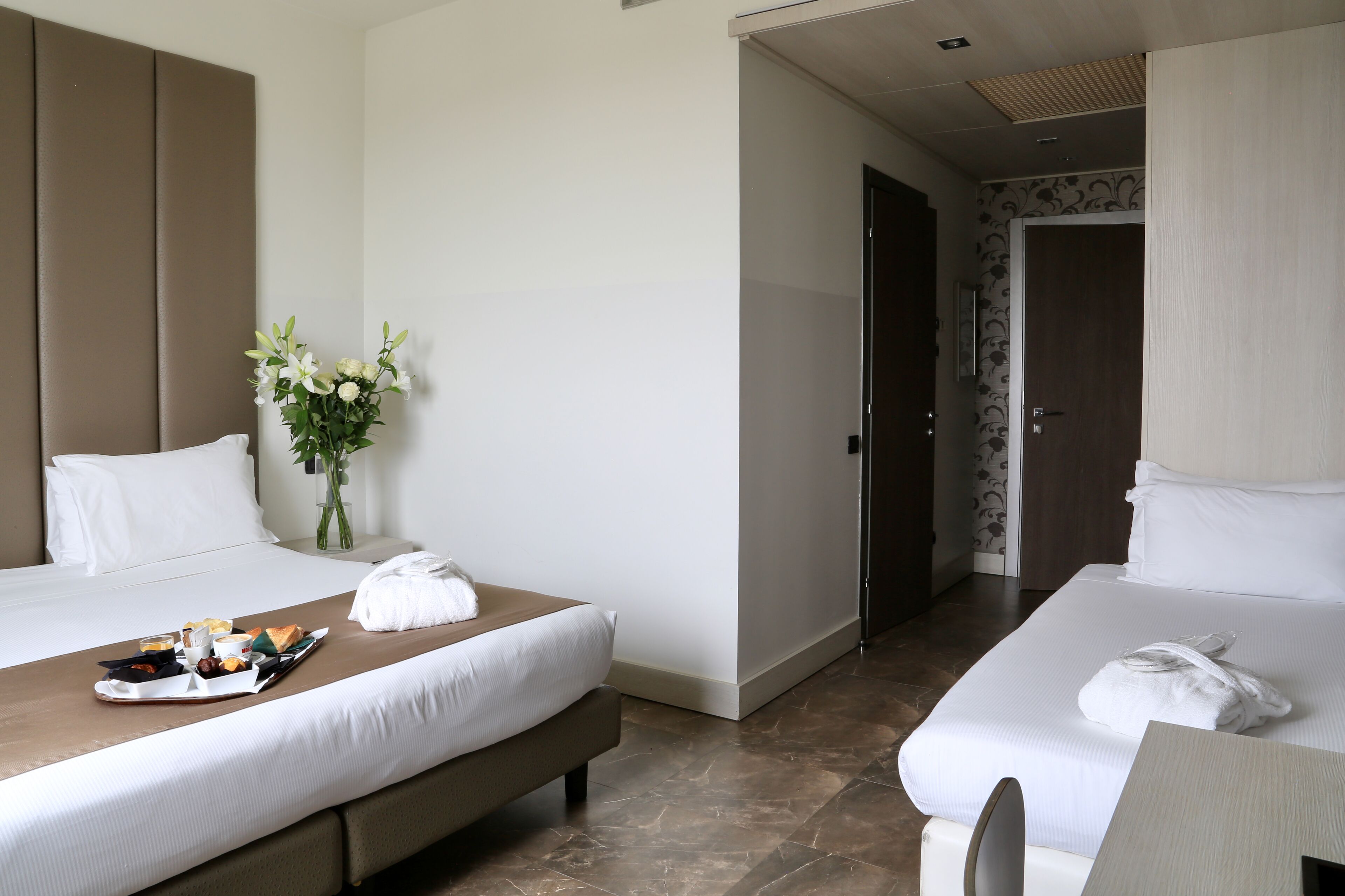 Habitación de hotel con dos camas, una bandeja de desayuno, un jarrón de flores y una puerta que da a otra habitación.