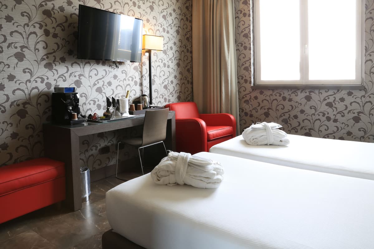 A hotel room with two beds, a desk with a coffee machine, a TV, and a red chair.