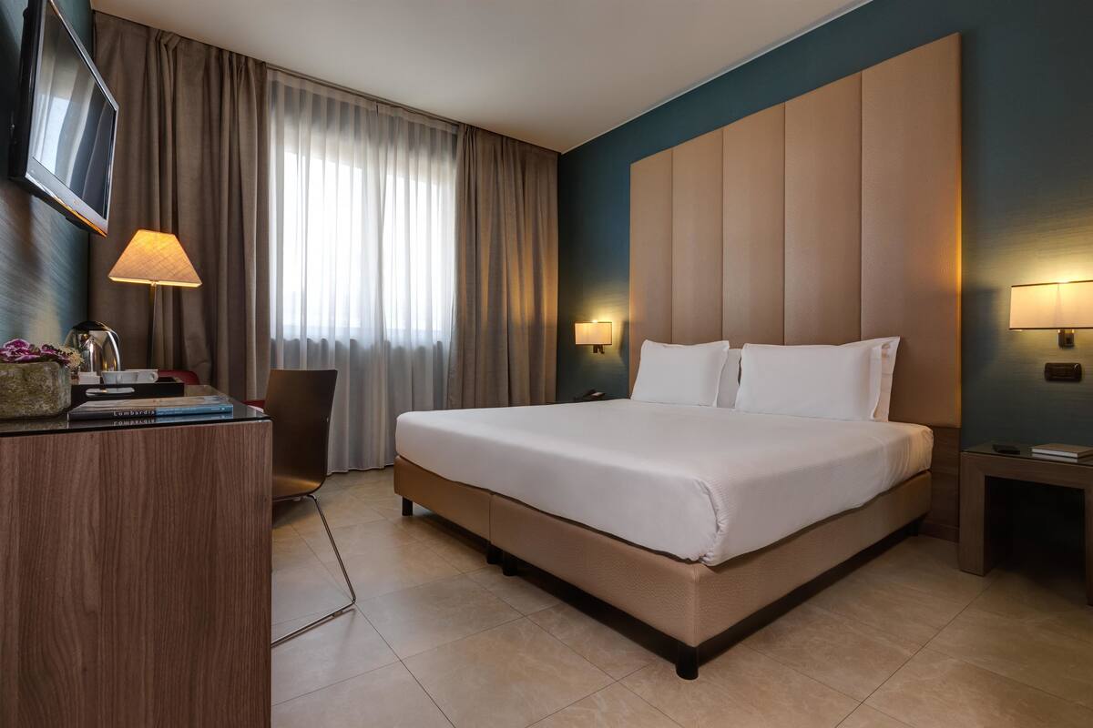 A hotel room with a large bed, a desk with a lamp, a TV, and a nightstand with a lamp.