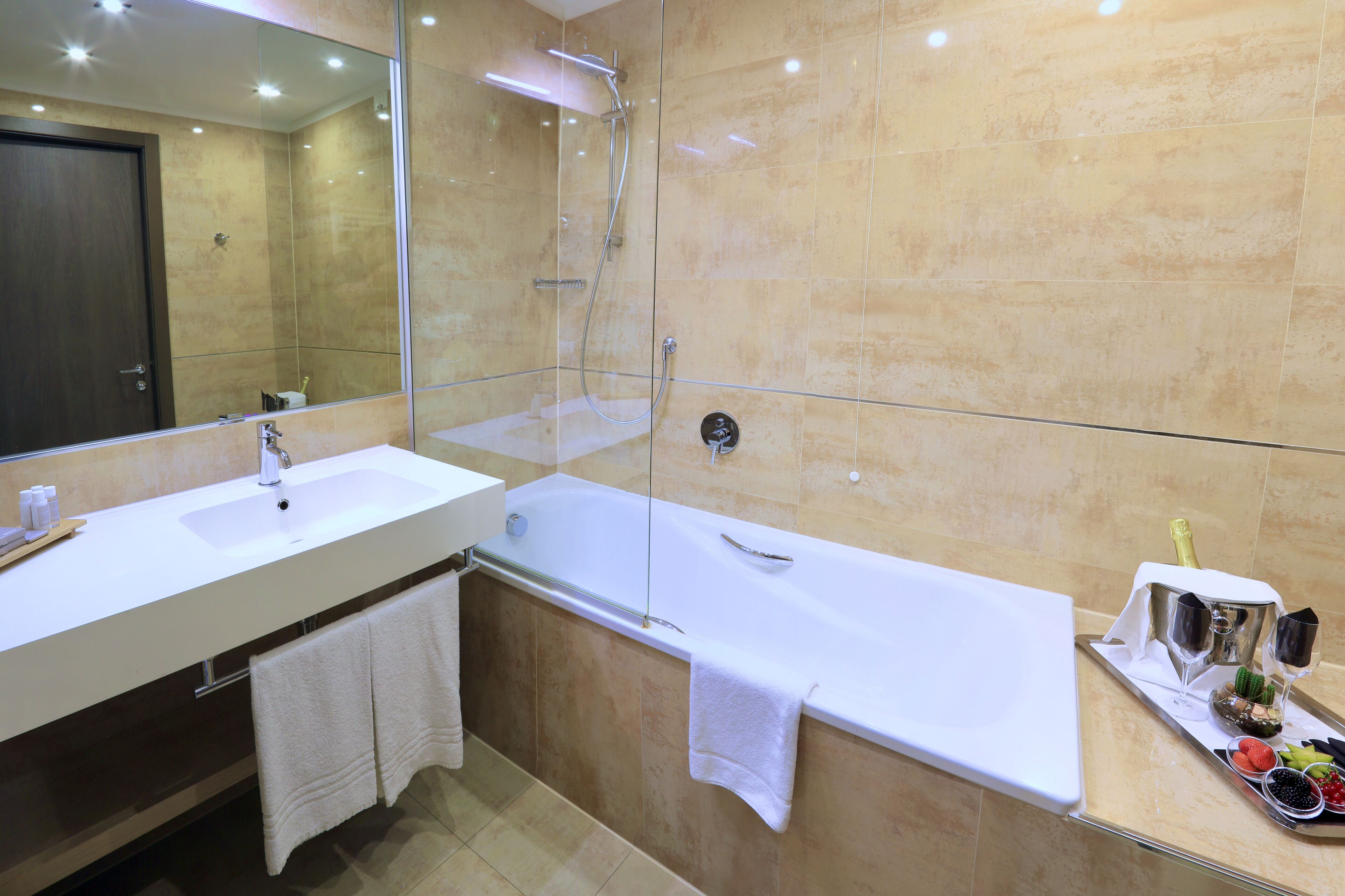 A modern bathroom with a large mirror, two sinks, a bathtub, and a tray with champagne glasses and fruits.