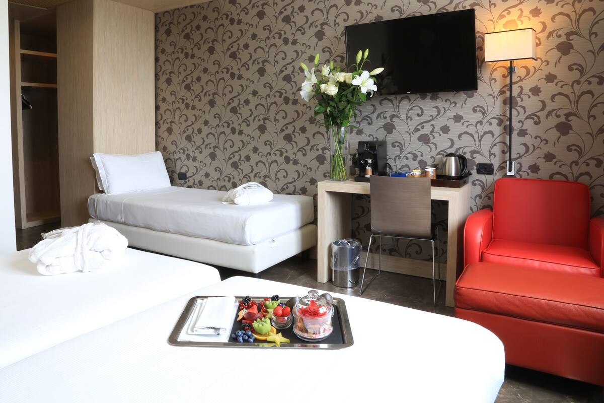 A hotel room with a bed, a desk, a red chair, and a tray with fruits and a coffee pot.