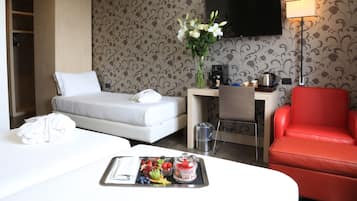 Ein Hotelzimmer mit Bett, Schreibtisch, einem roten Sessel und einer Ablage mit Früchten und einer Kaffeemaschine.