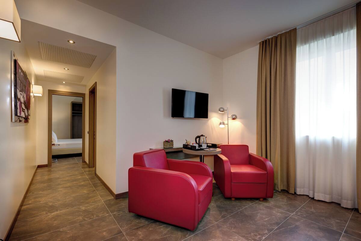 A hotel room with a modern design, featuring a seating area with red chairs, a flat-screen TV mounted on the wall, and a small table with a lamp.