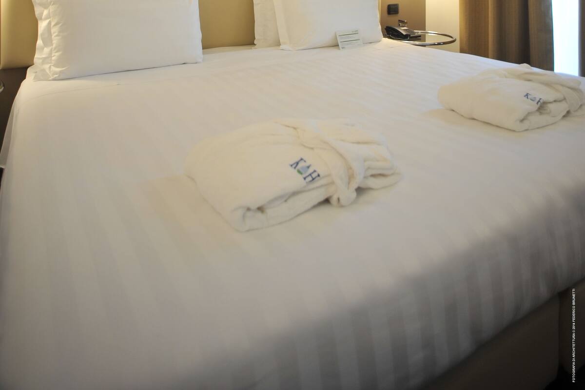 A hotel room with a large bed, two pillows, a bedside table with a lamp, and a window with curtains.
