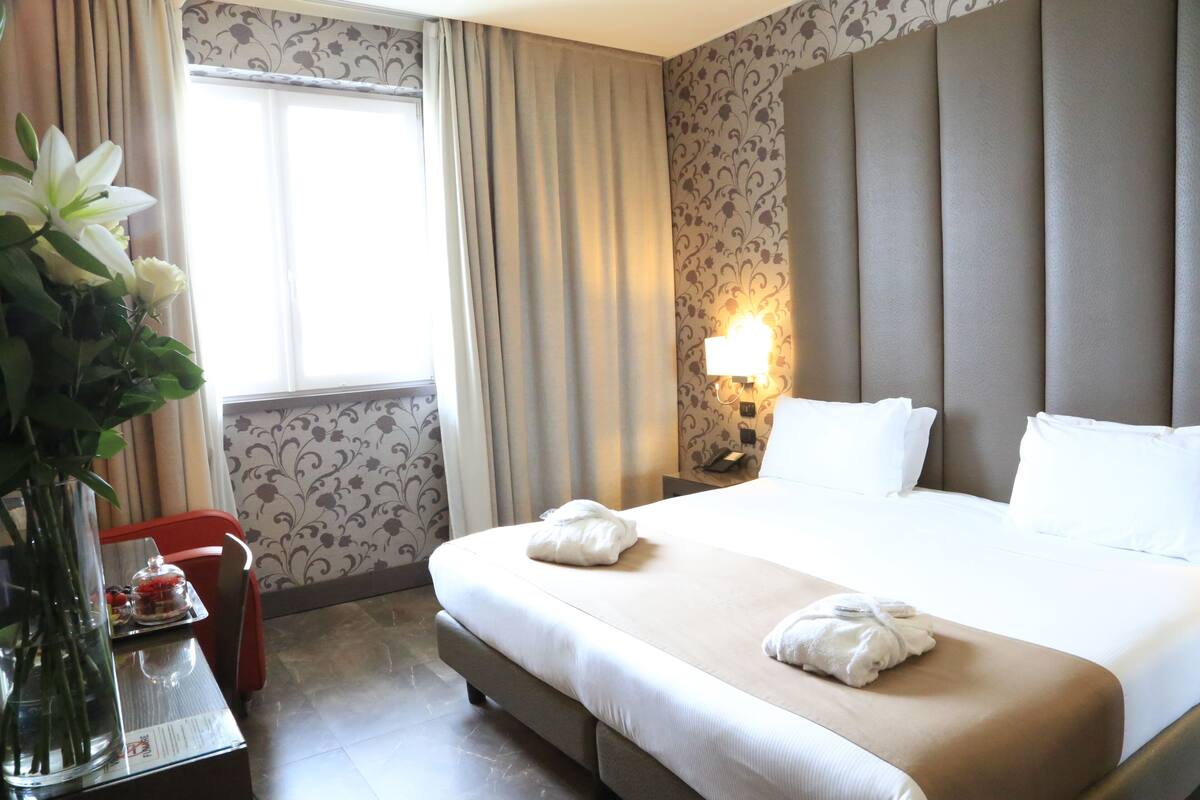 A hotel room with a large bed, a bedside table with a lamp, a vase with flowers, and a window with curtains.