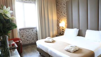 A hotel room with a large bed, a bedside table with a lamp, a vase with flowers, and a window with curtains.