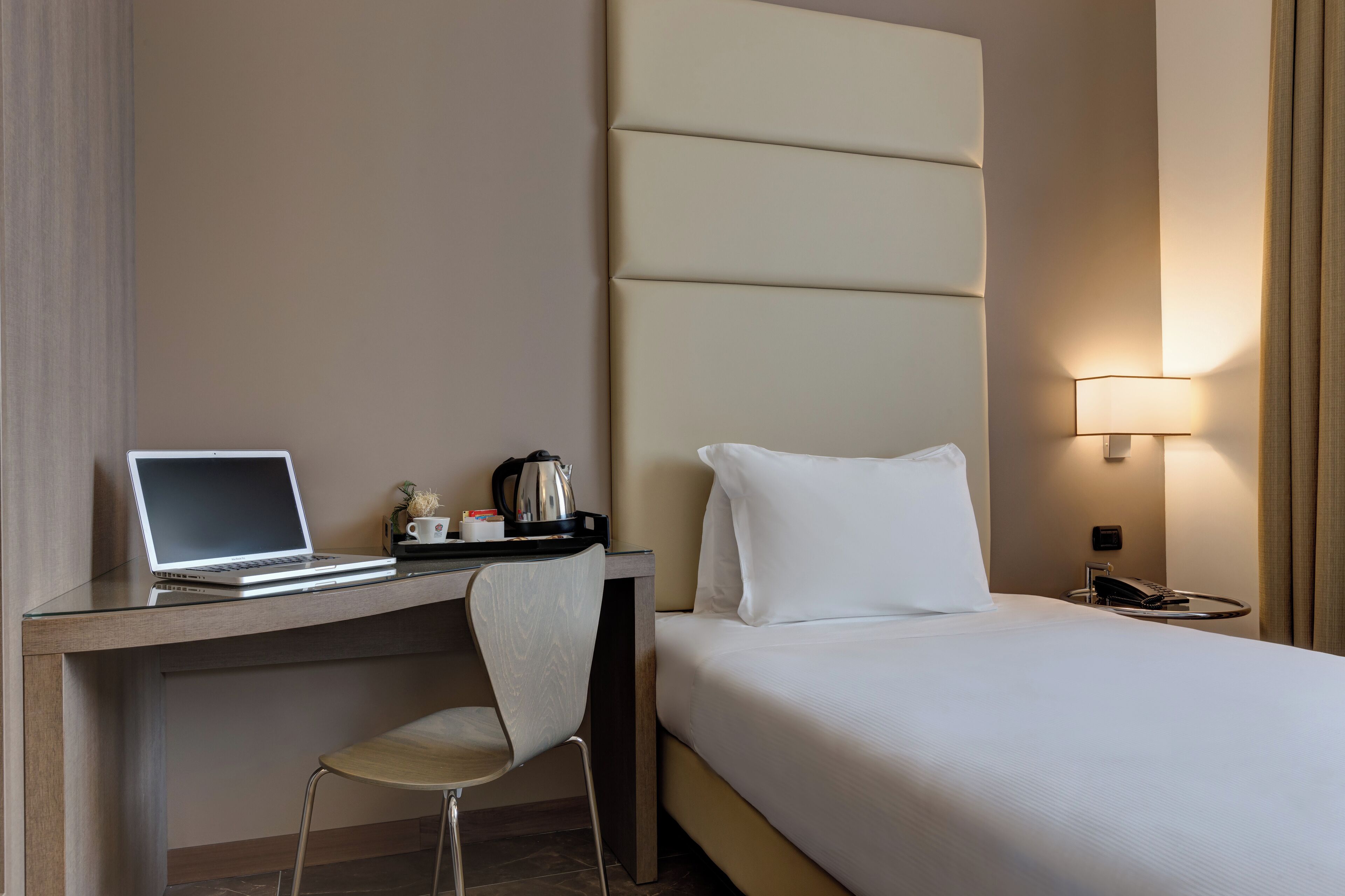 Habitación de hotel con cama, escritorio con computadora portátil, silla, mesita de noche con lámpara y otro buró.