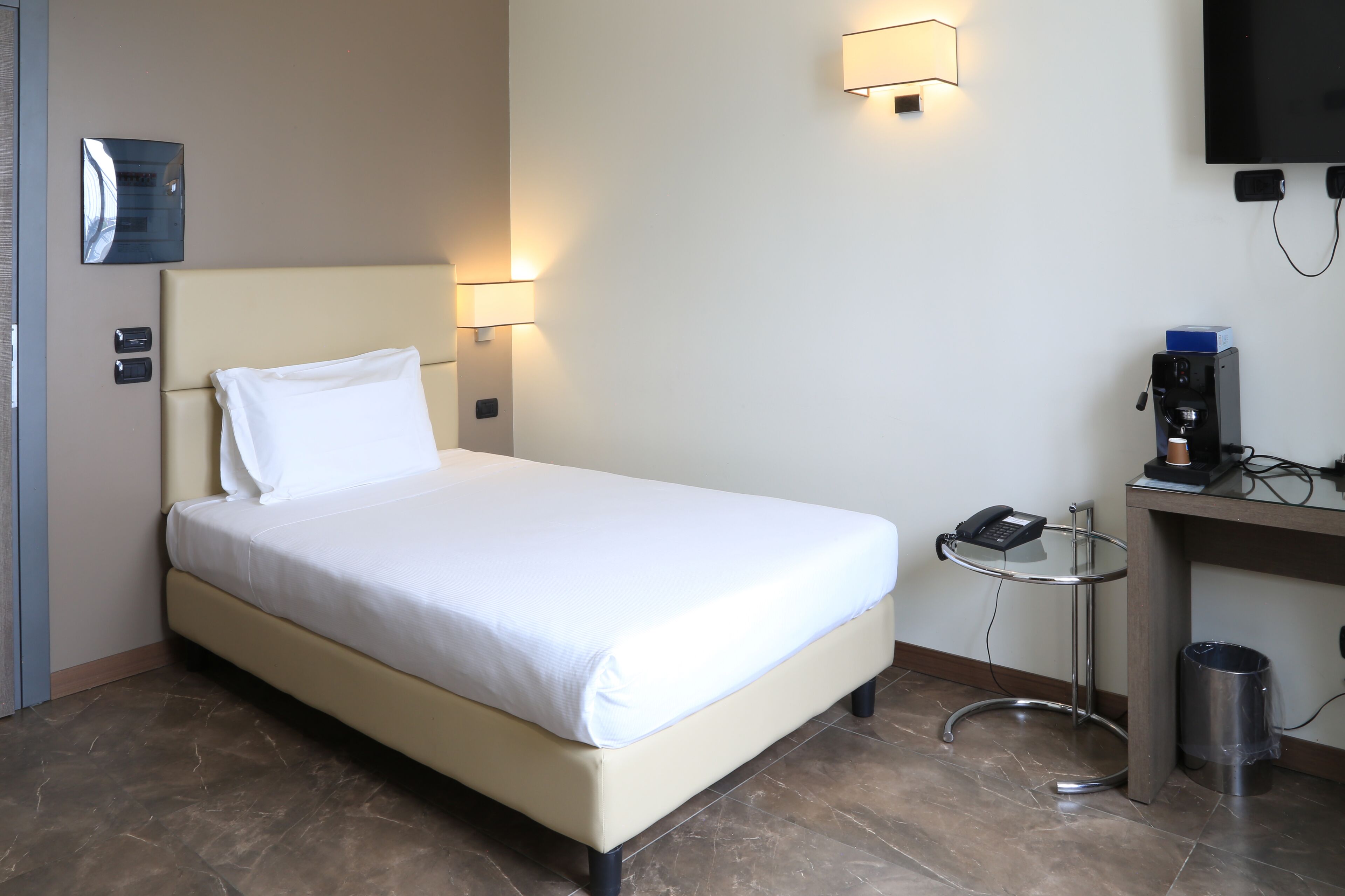 Una habitación de hotel con cama, lámparas de noche, televisor, cafetera y cubo de basura.