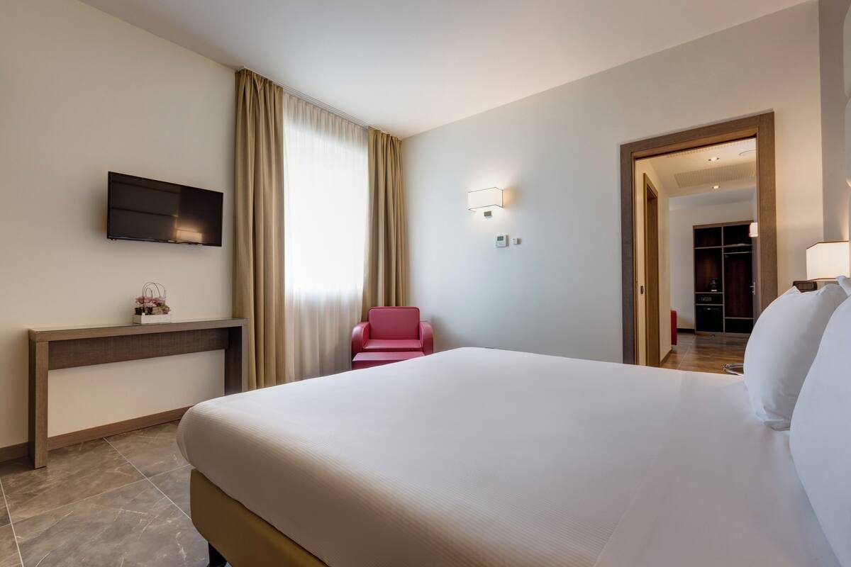 A hotel room with a large bed, a TV mounted on the wall, a desk with a chair, and a wardrobe in the background.