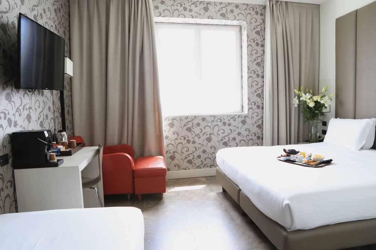 A hotel room with a bed, a desk with a coffee maker, a TV, and a window with curtains.