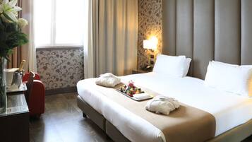 Een hotelkamer met een groot bed, een nachtkastje met een lamp, een schaal met fruit en een rode koffer.
