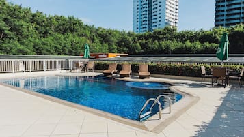 Una piscina con sillas de descanso y sombrillas, rodeada por un suelo de baldosas y follaje.